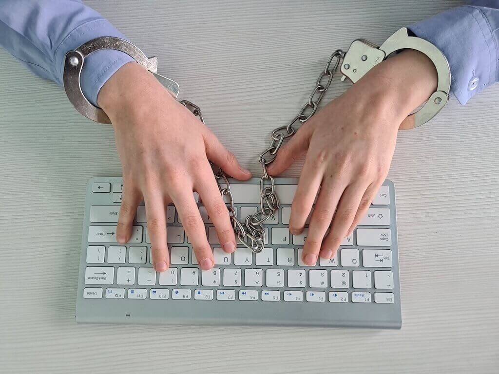 Hands in handcuffs typing on a keyboard illustrating ethical issues related to digital restrictions and online freedom